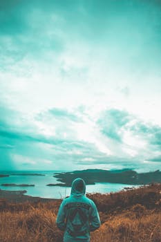 A person in a hoodie stands on a hilltop, gazing at a scenic ocean view under moody skies.