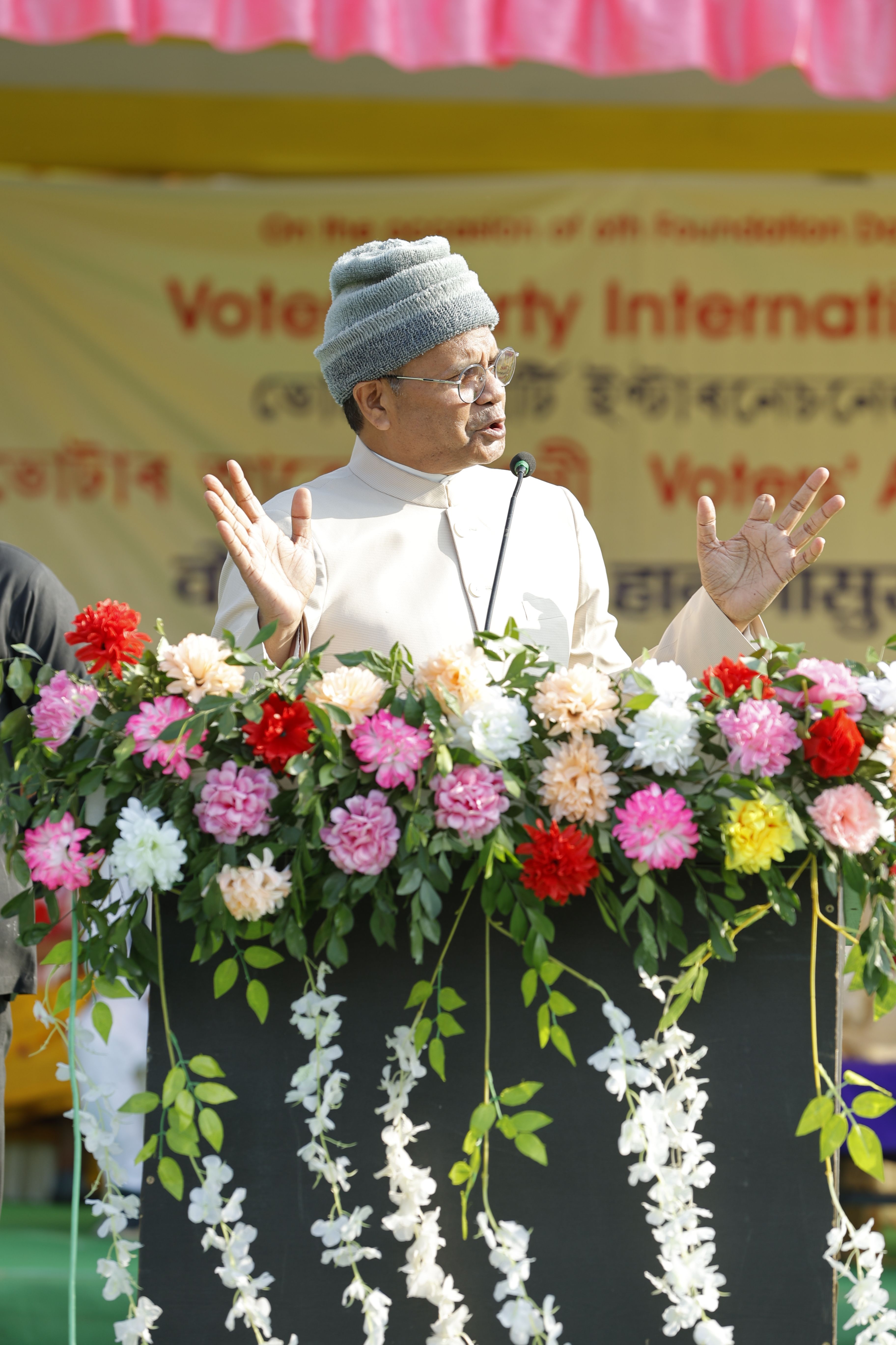 A man wearing a cap delivers a speech at a podium adorned with colorful flowers, captured during an outdoor event.