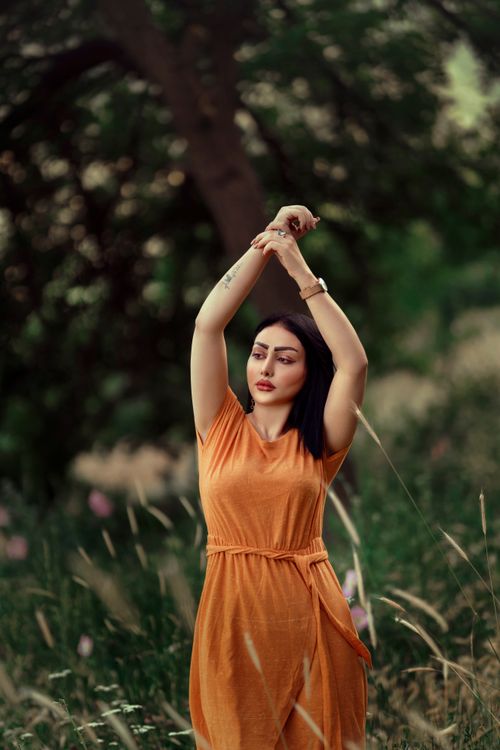 Free Woman in an orange dress poses gracefully in a lush green meadow, arms raised. Stock Photo