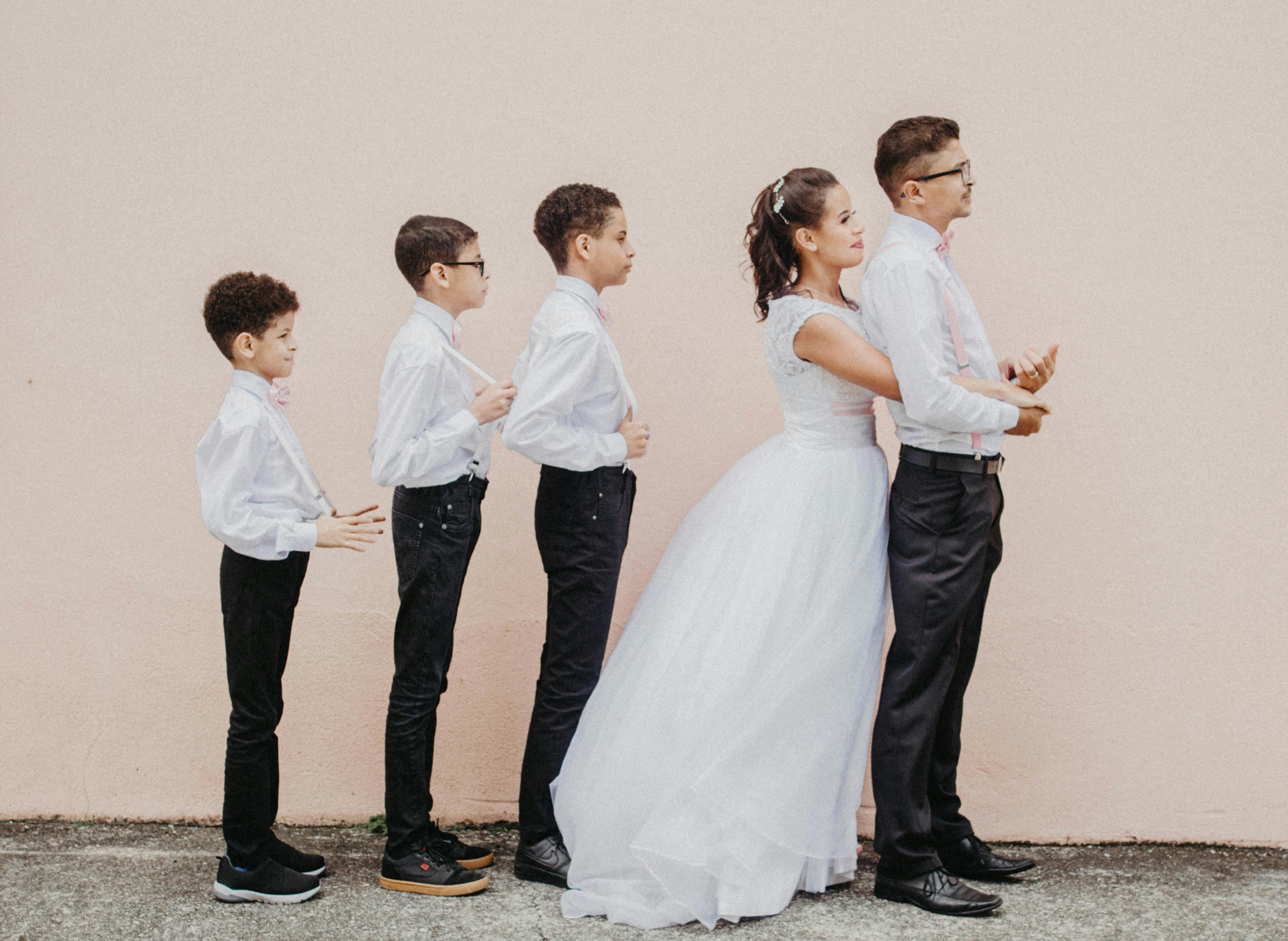 Charming family wedding portrait with bride, groom, and children standing in line against a pink wall.