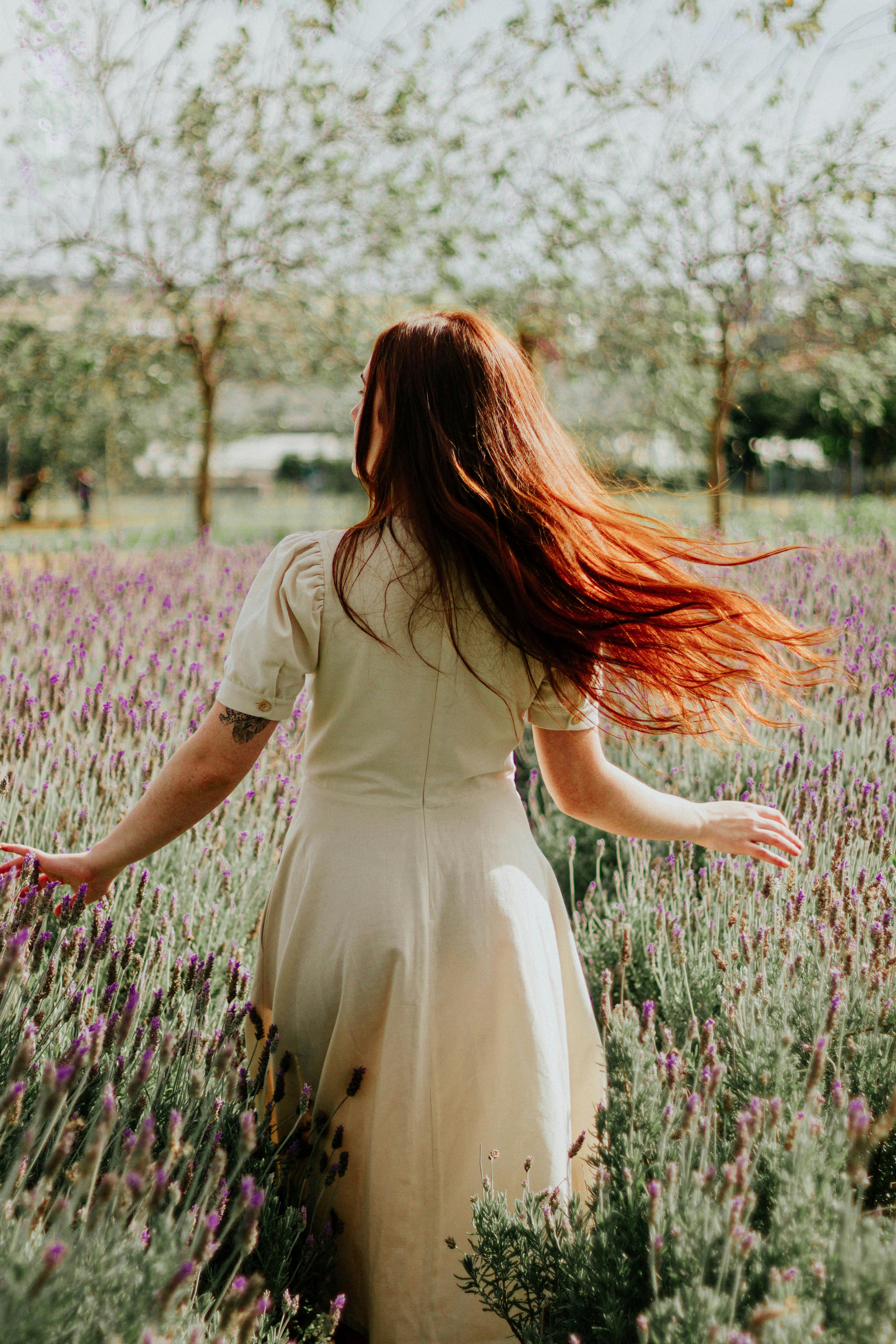 A woman with red hair walks gracefully through a vibrant lavender field under a sunny sky.