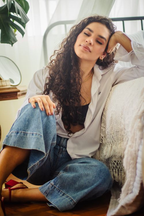 Free A woman with long curly hair relaxes with eyes closed, sitting indoors in a cozy setting wearing casual fashion. Stock Photo