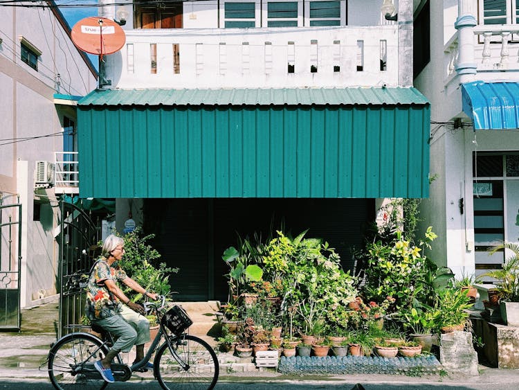 Old Woman Riding Bike Near Plants Shop