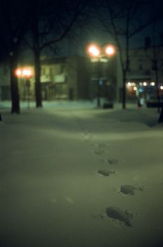 Snow-covered city street at night with footprints leading into the distance, illuminated by streetlights.