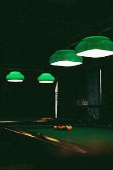 Dimly lit billiards room with a pool table and green hanging lamps creating a moody atmosphere.