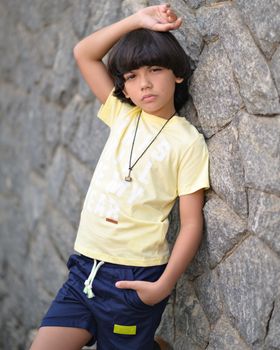 A stylish young boy with black hair poses confidently against a stone wall in casual attire.