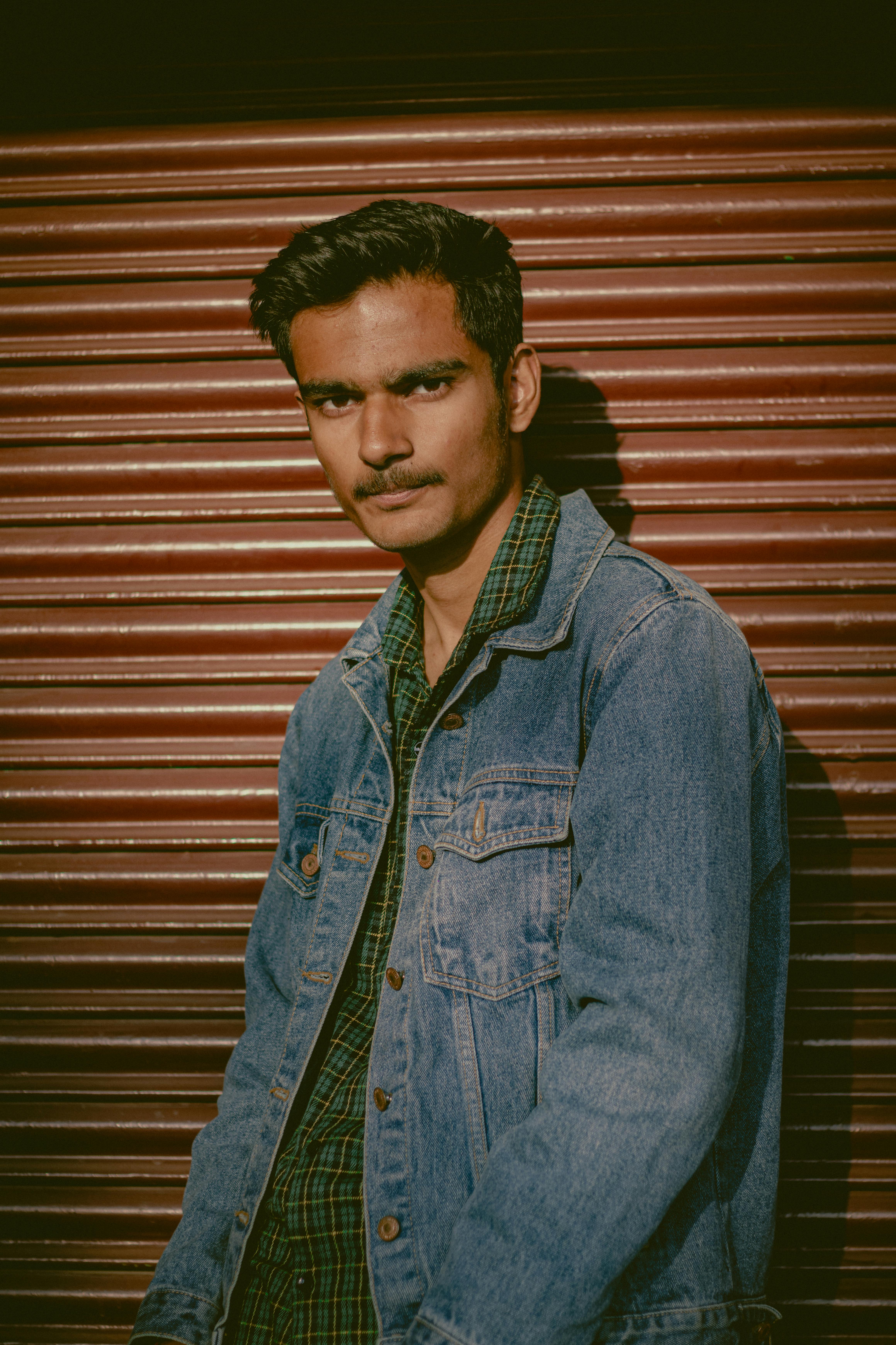 Portrait of a young South Asian man wearing a denim jacket in front of a rolling door.