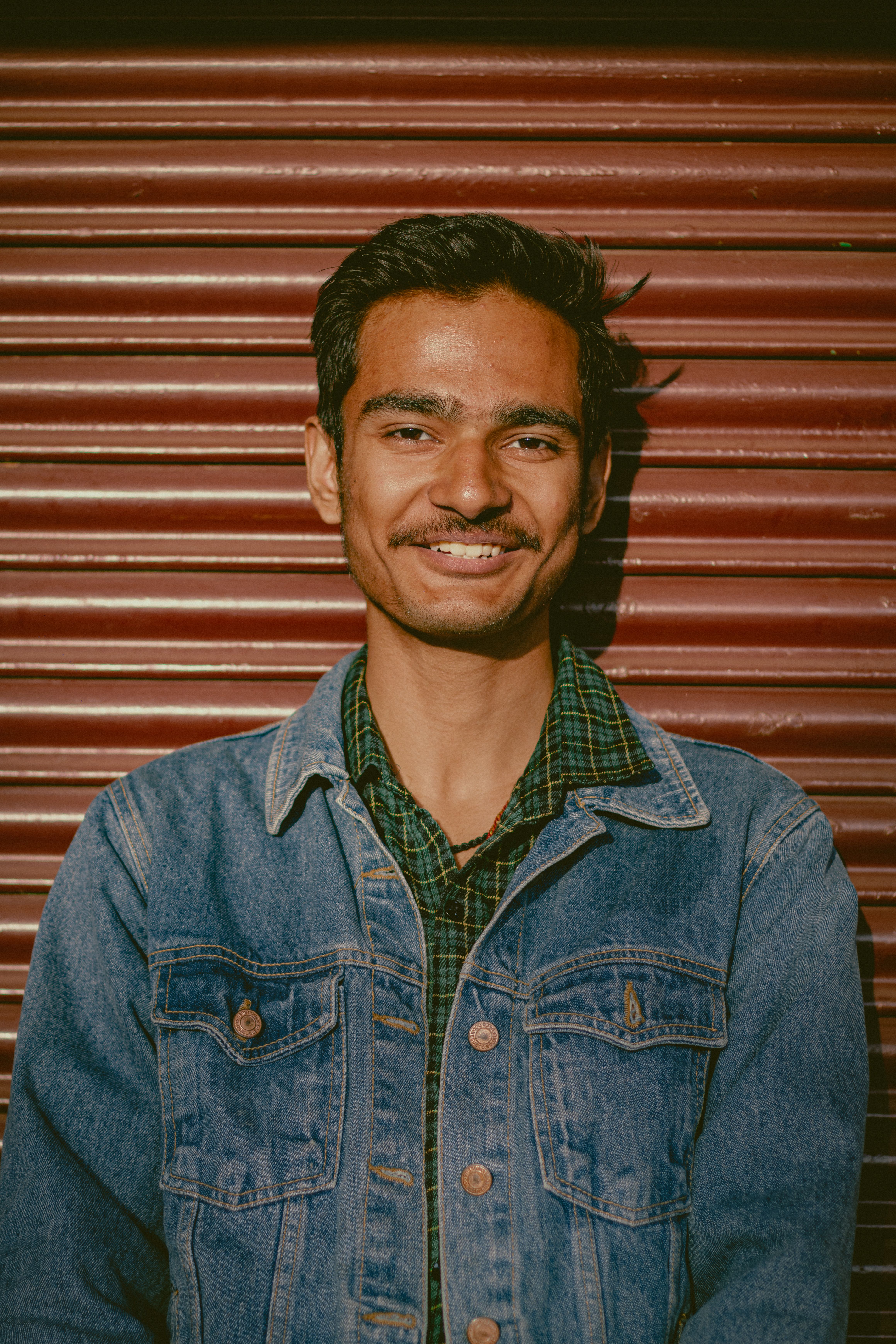 Smiling young man in denim jacket poses confidently against a metal shutter backdrop.