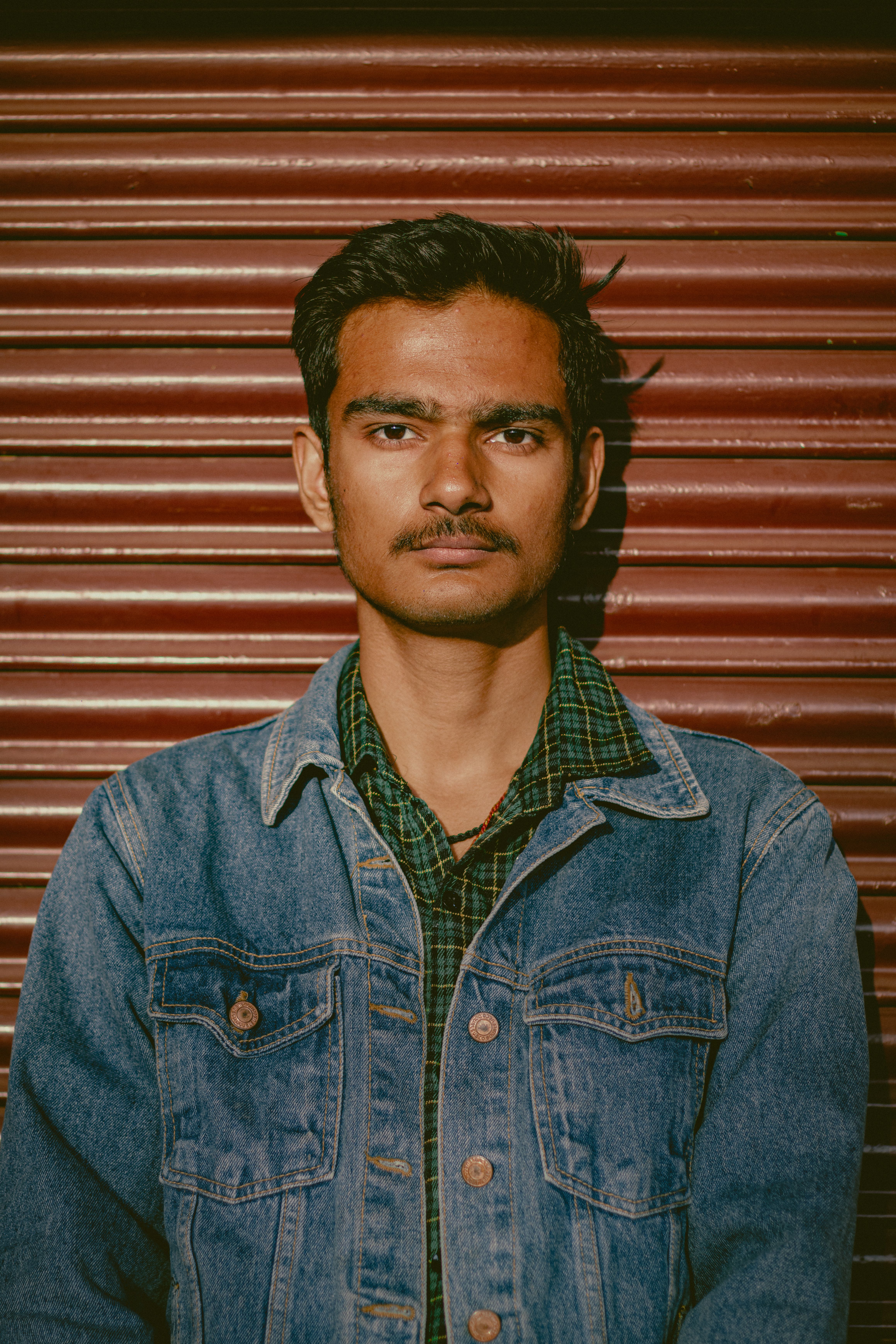Portrait of a young man in denim and green check shirt standing against a rolling door, under natural lighting.