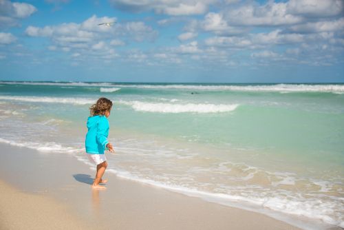 Free A playful child in blue runs along a vibrant beach with waves and clear skies. Stock Photo