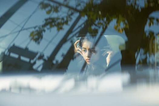 A thoughtful woman with headphones seen through a car window reflection.