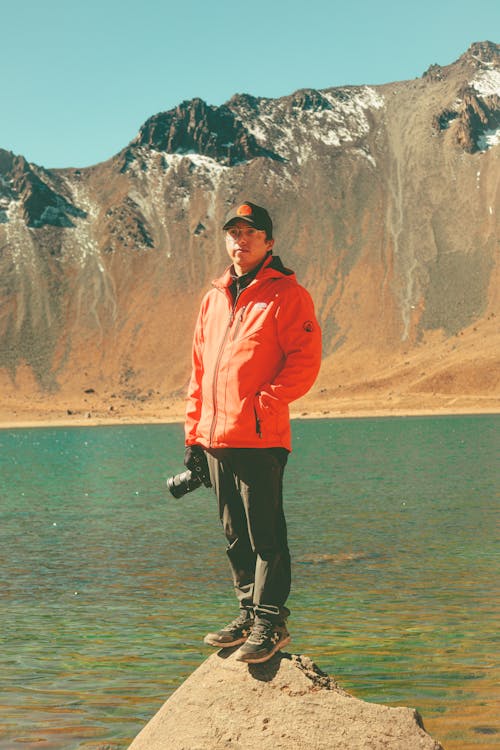 Man Wearing a Red Jacket, Standing with a Camera by a Lake in a Mountain Landscape