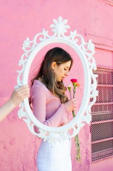 Young woman viewed through an ornate mirror holding flowers with vibrant pink background wall.