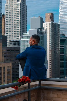 A man in a blue suit sits on a balcony with roses, gazing at an urban skyline.