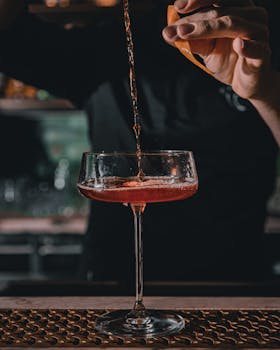 A bartender pours a refreshing cocktail garnished with orange peel into a glass at a stylish bar.