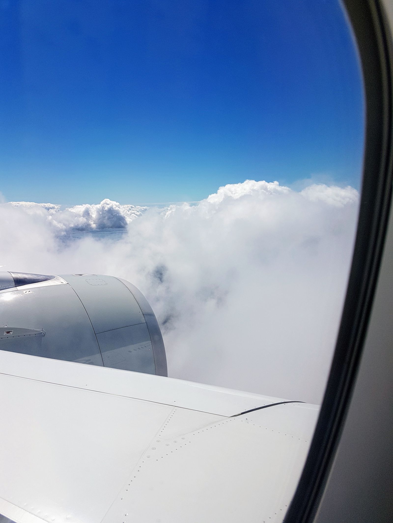 View from an airplane window seat over the wing, clouds and blue sky beyond
