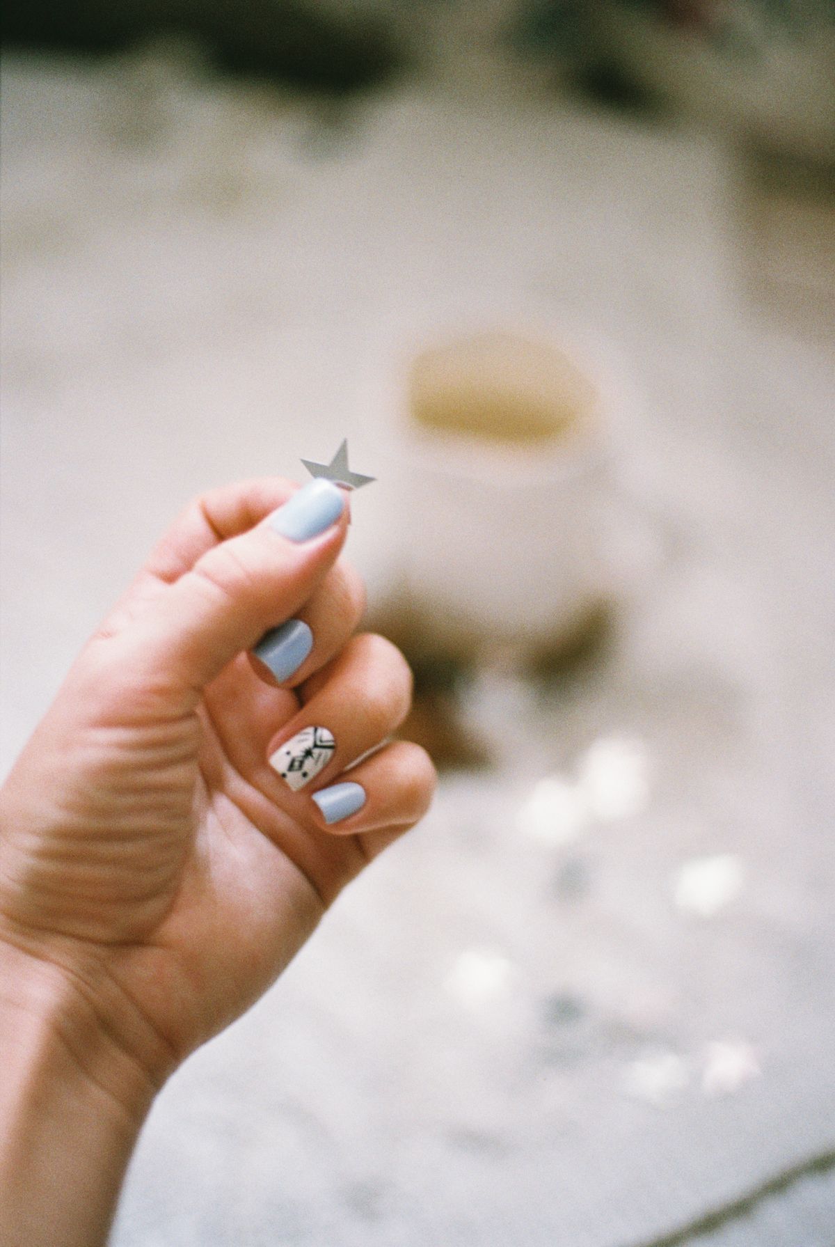 A close-up of a hand with blue painted nails holding a silver star confetti piece over a blurred festive background.