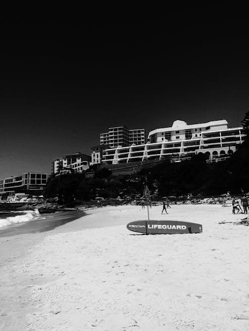 Black and White Photography of a Surfboard on a Beach and a Tourist Resort