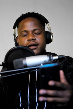 A close-up portrait of a man singing into a microphone, wearing headphones, and holding a cellphone.