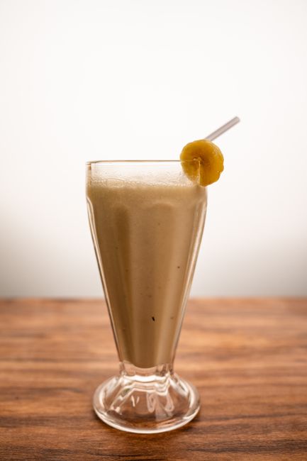 A thick banana peanut butter protein smoothie in a tall glass, topped with banana slices and a drizzle of peanut butter, with a metal straw and scattered peanuts on the counter.