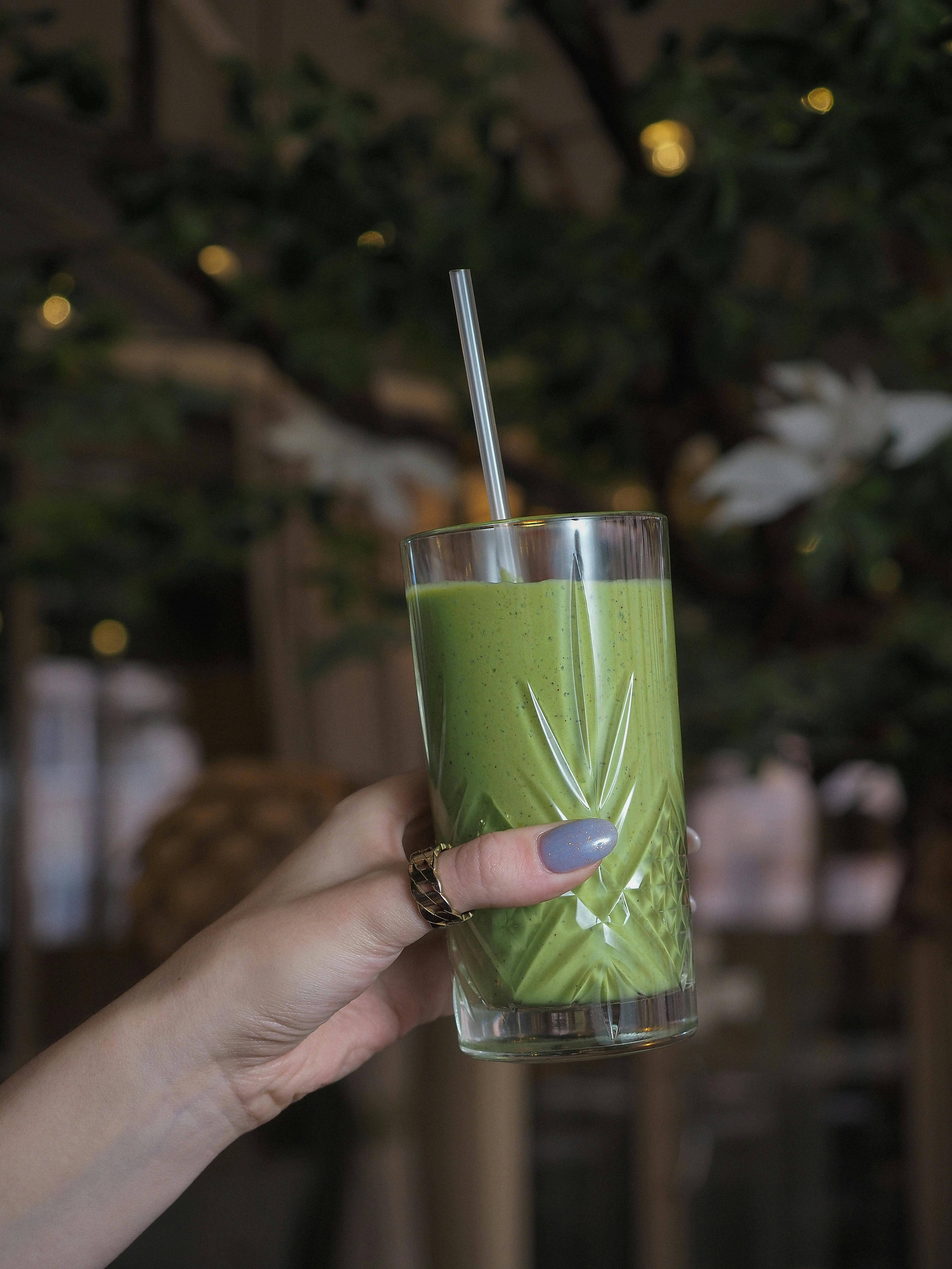 Close-up of a green smoothie in a glass held by a hand indoors.