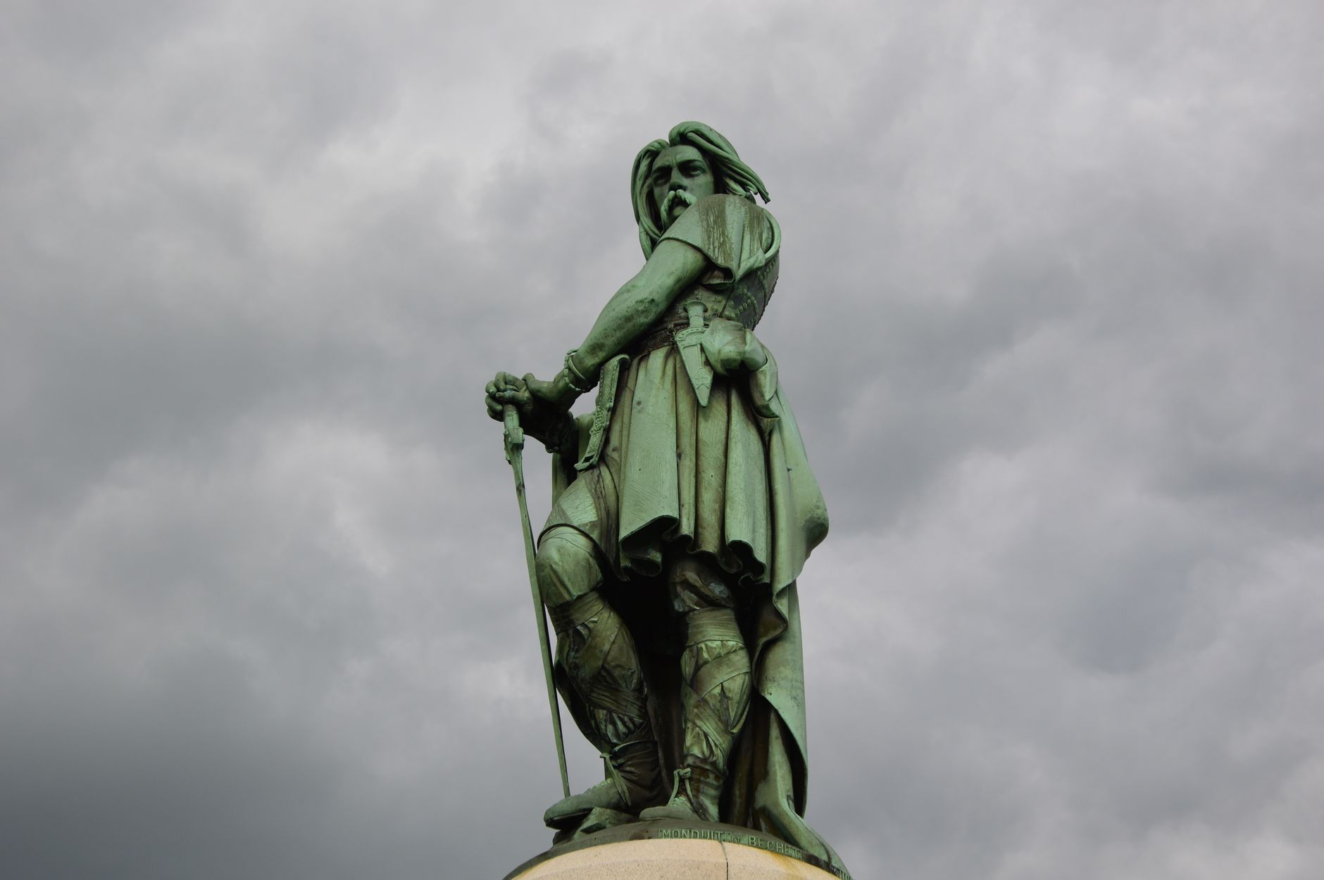 A striking bronze warrior statue stands tall against an ominous overcast sky, captured in a low angle shot.