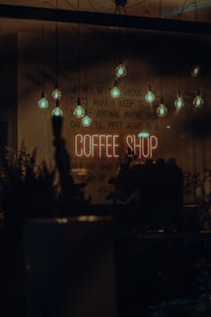 Warm and inviting coffee shop interior with glowing neon sign and hanging light bulbs, creating a cozy ambiance at night.
