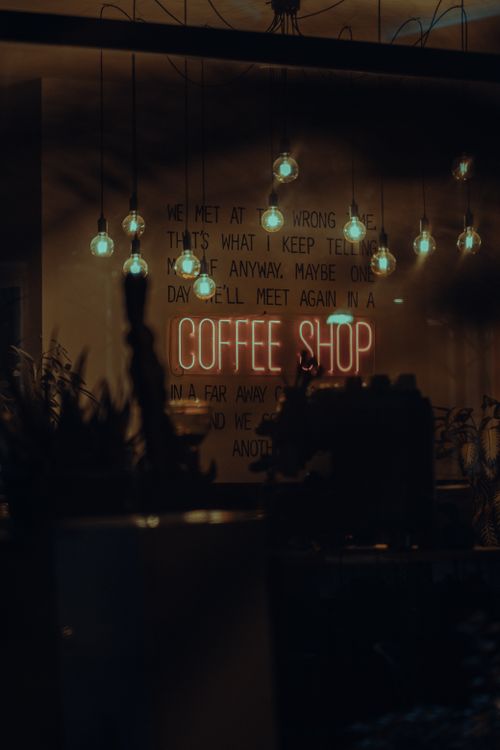 Free Warm and inviting coffee shop interior with glowing neon sign and hanging light bulbs, creating a cozy ambiance at night. Stock Photo