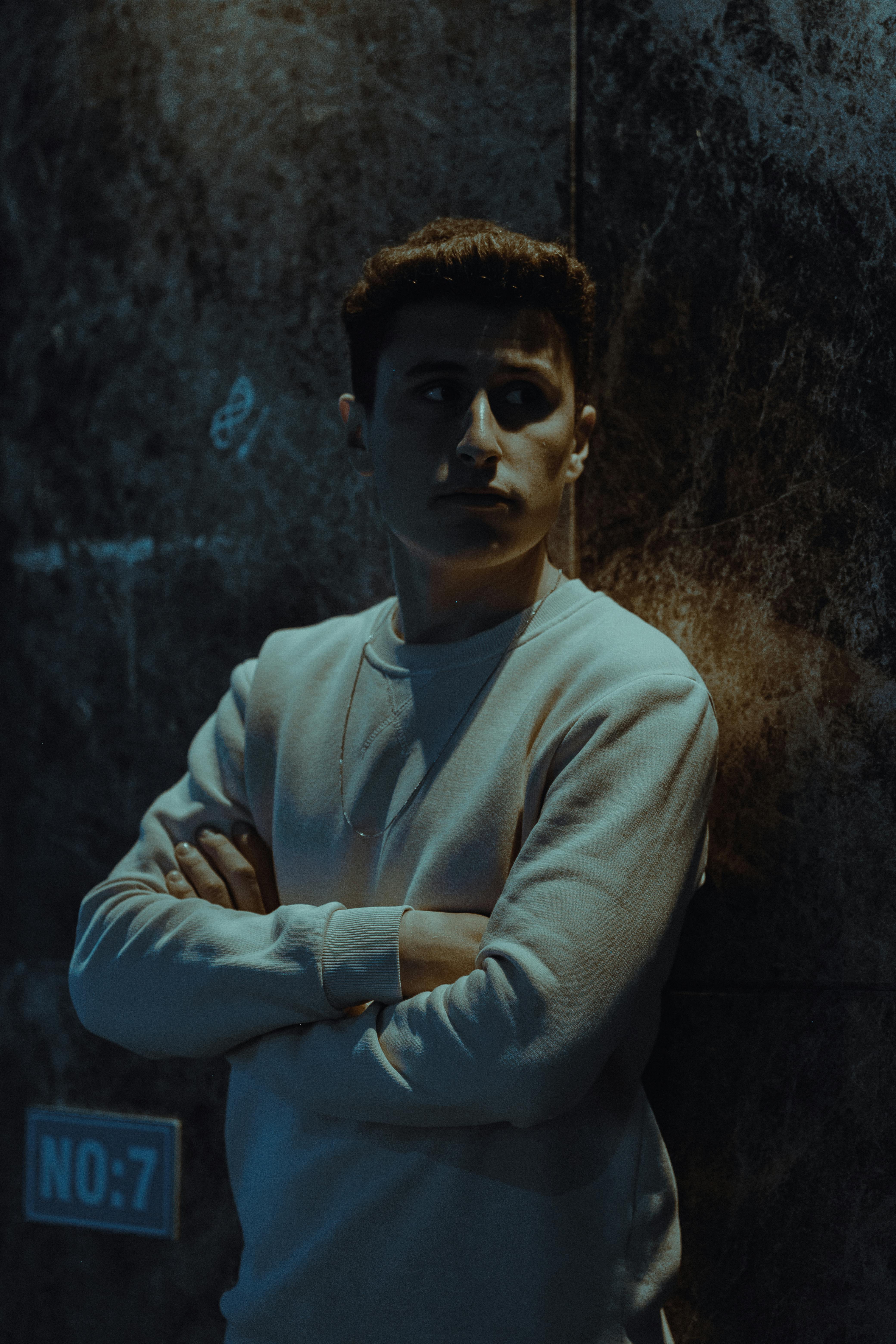 A man with arms crossed stands against a textured wall at night, exuding a moody urban vibe.