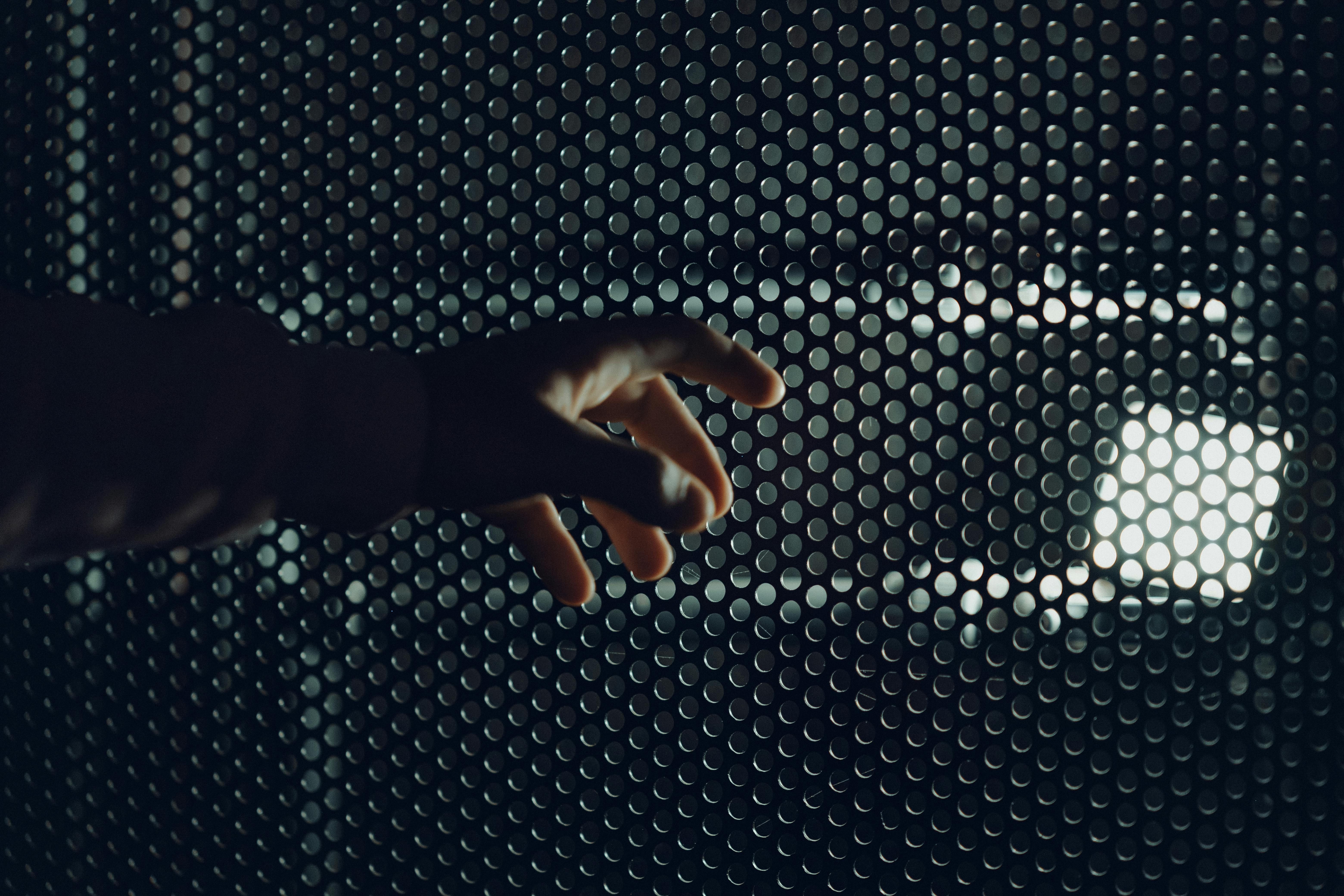A human hand reaches towards a light beyond a perforated metal grid, creating a dramatic effect.