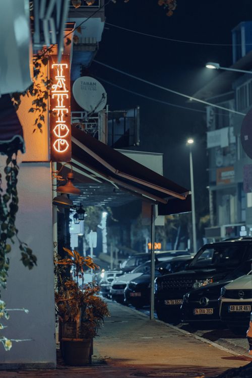 Free A bustling nighttime street scene featuring a glowing neon tattoo sign and parked cars. Stock Photo