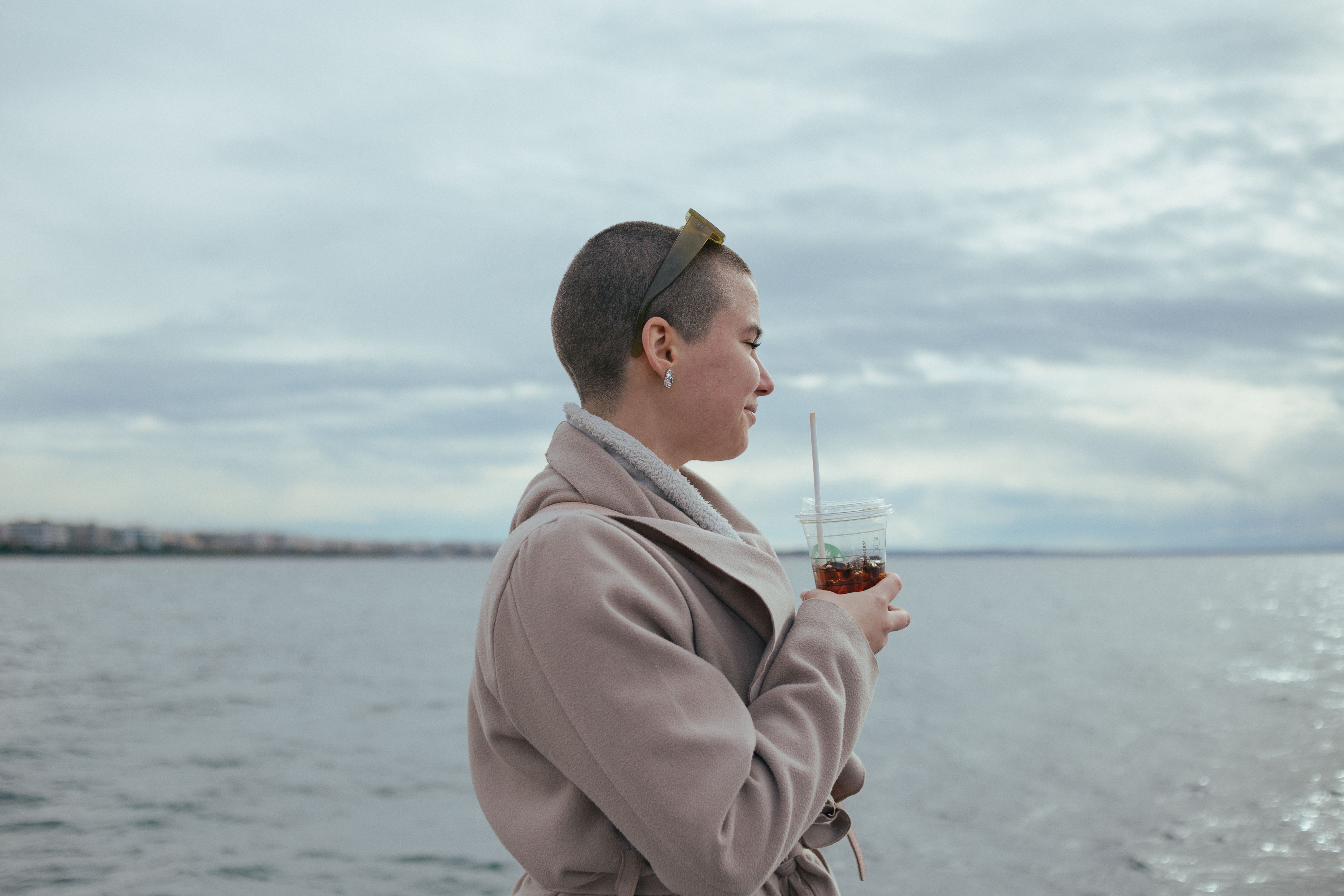Woman with short hair holding a drink, smiling by a calm sea under cloudy skies.