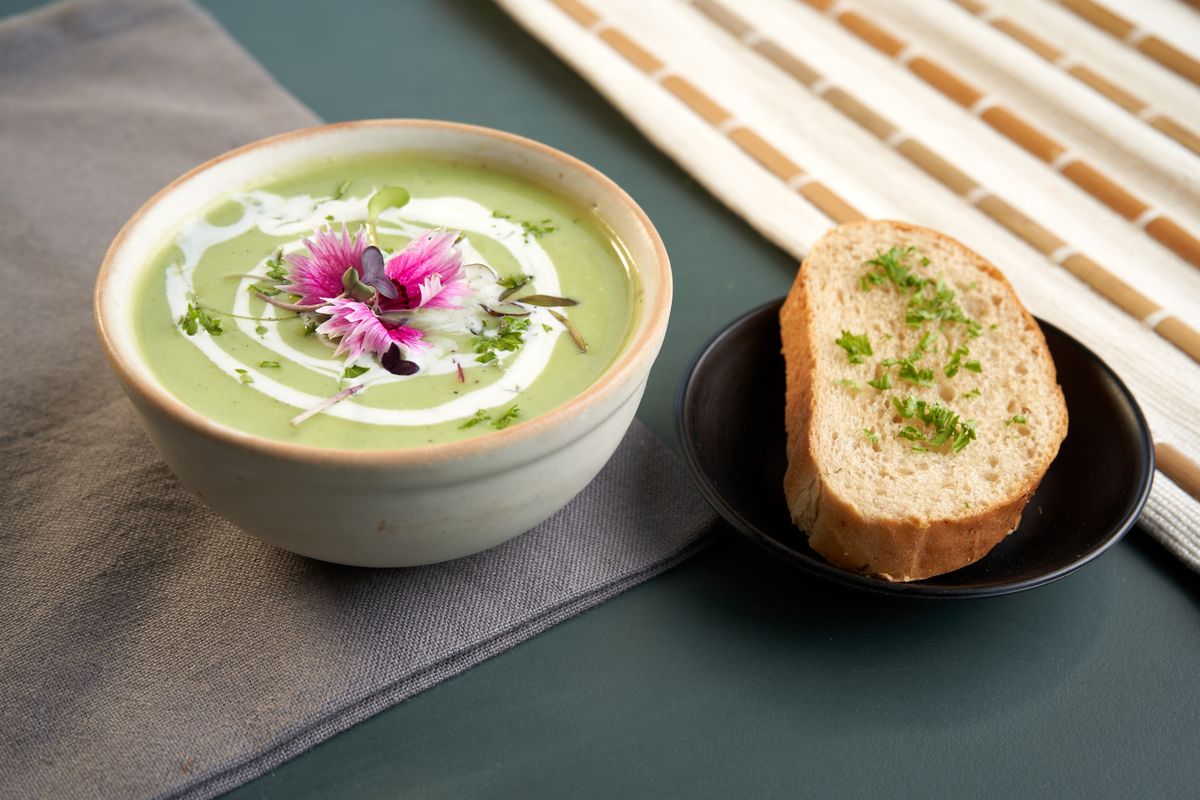 Pale green zucchini soup with cream swirl, pink edible flower, beside herb-topped bread slice