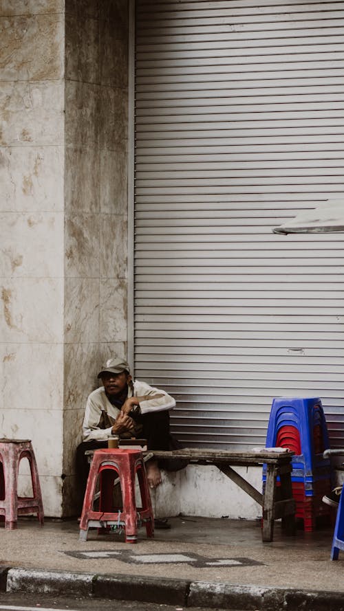 Man Sitting on a Sidewalk, in the Corner of a Building in City 