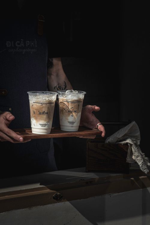 Free Barista presenting two iced lattes on a wooden tray in a dimly lit cafe setting. Stock Photo