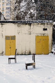 Abandoned chairs in a snowy urban area with vibrant yellow doors.
