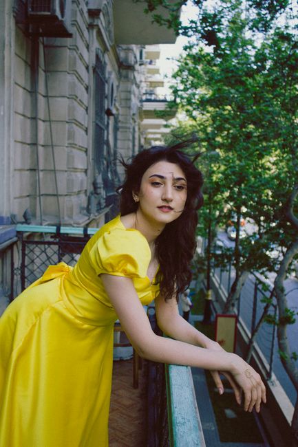 An elegant woman in a yellow dress leaning on a balcony with cityscape background.
