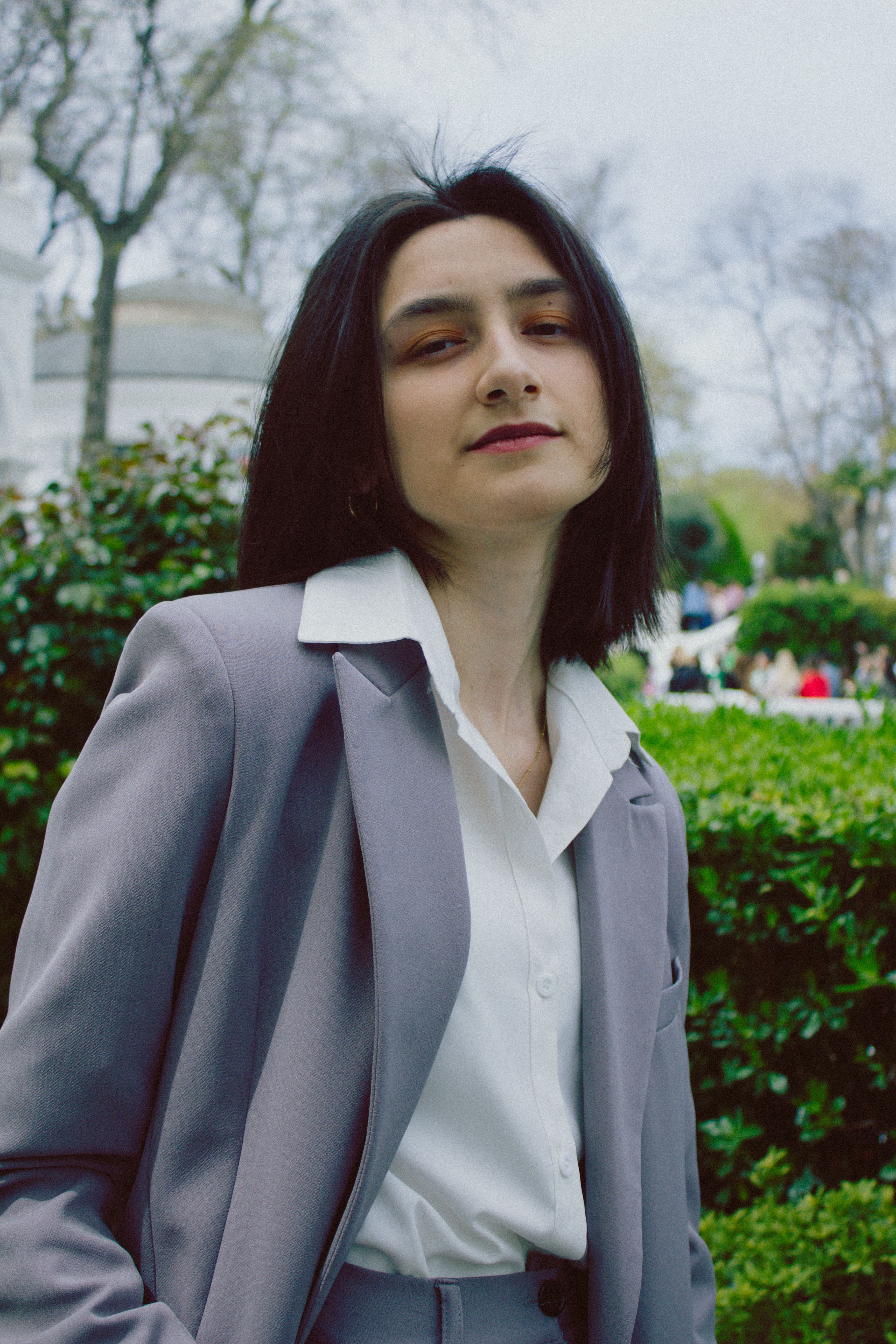 A confident businesswoman wearing formal attire, posing outdoors with greenery in the background.