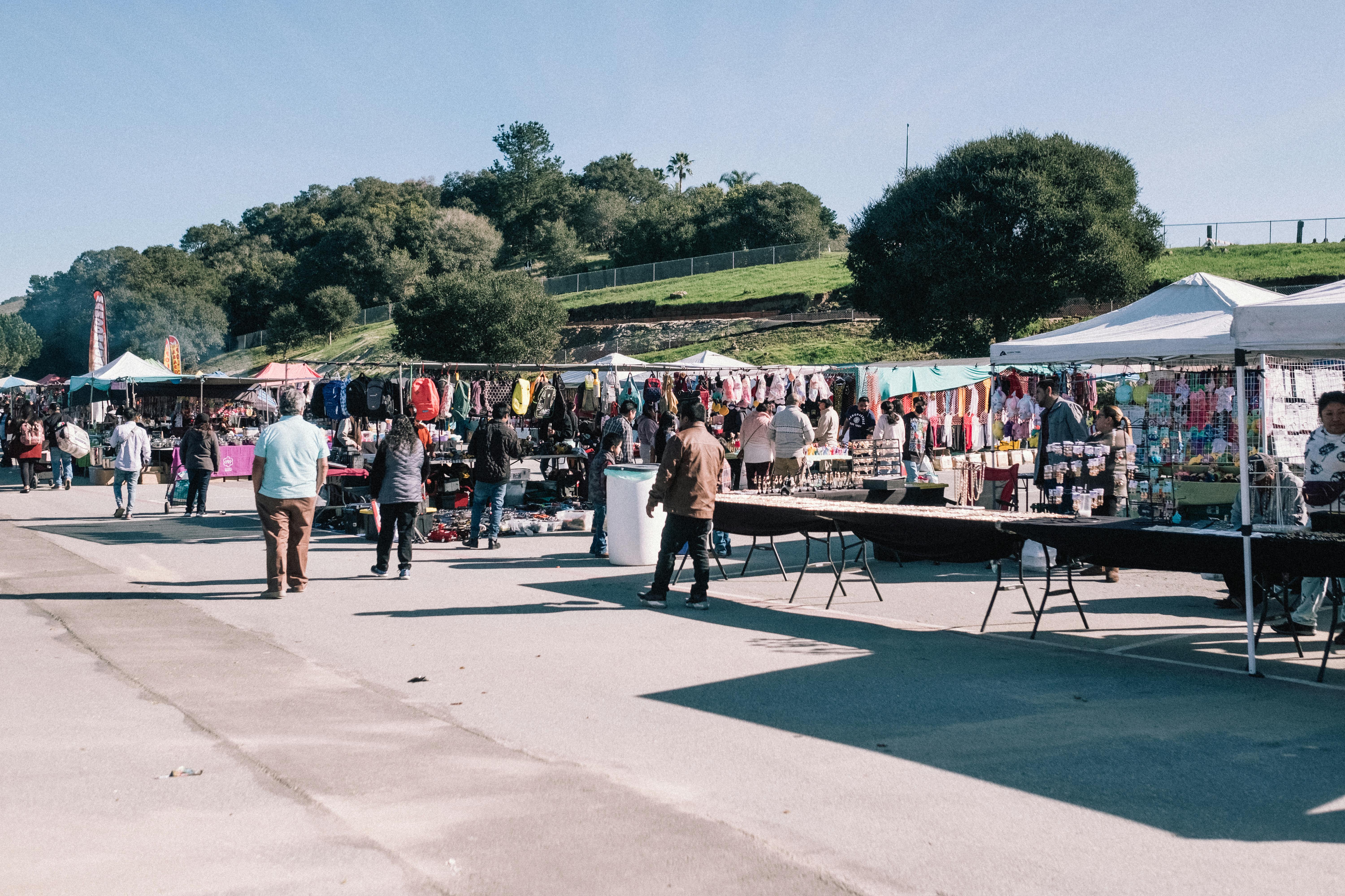 Busy outdoor market scene with people shopping under tents on a sunny day.