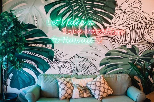 Free A cozy lounge with tropical leaves and neon sign saying 'Let's take a Boba Vacation to Hawaii'. Stock Photo