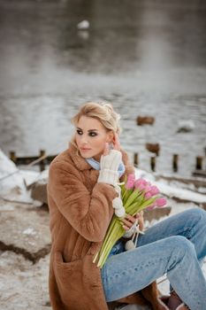 Caucasian woman in winter attire holding pink tulips by a snowy lake.