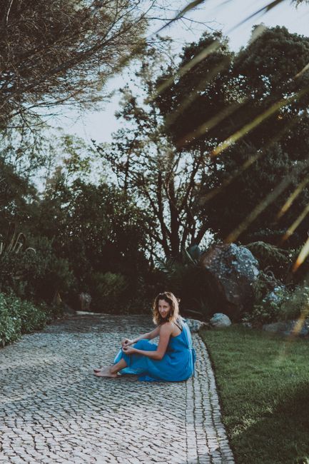 A woman in a blue dress sitting peacefully on a cobblestone path in a lush green park setting, enjoying summer.