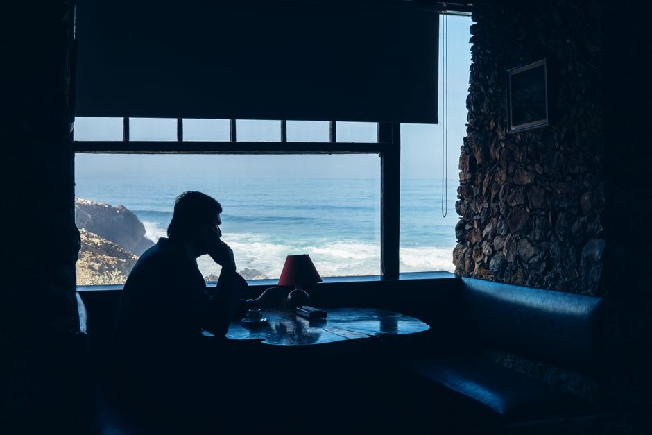 A contemplative man sits at a seaside restaurant, enjoying ocean views through a window.