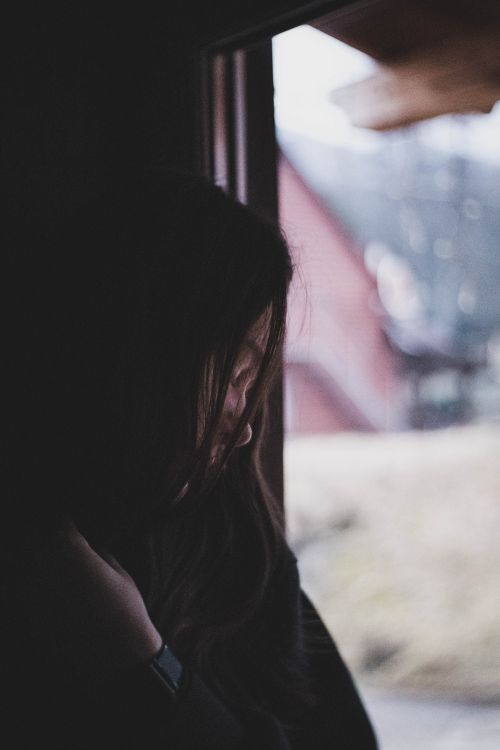 Free A woman gazes thoughtfully out of a window, framed by soft shadows and muted lighting. Stock Photo