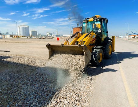 A yellow excavator unloading pebbles on a road at an industrial construction site under a clear blue sky.