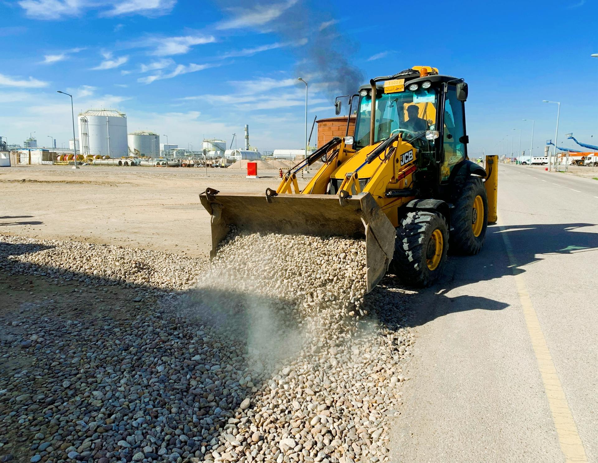 A yellow excavator unloading pebbles on a road at an industrial construction site under a clear blue sky.