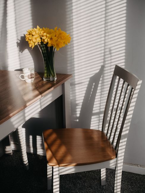 A warm still life scene with daffodils in a vase, set on a wooden table with a chair and soft light.
