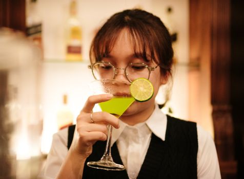 A woman with glasses holds a lime-green cocktail in a bar setting.