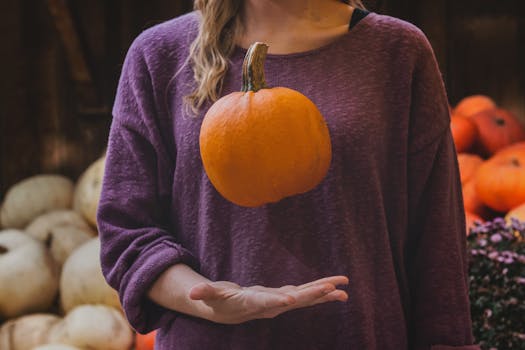 A woman in a sweater holds a hovering pumpkin, symbolizing fall's harvest season.