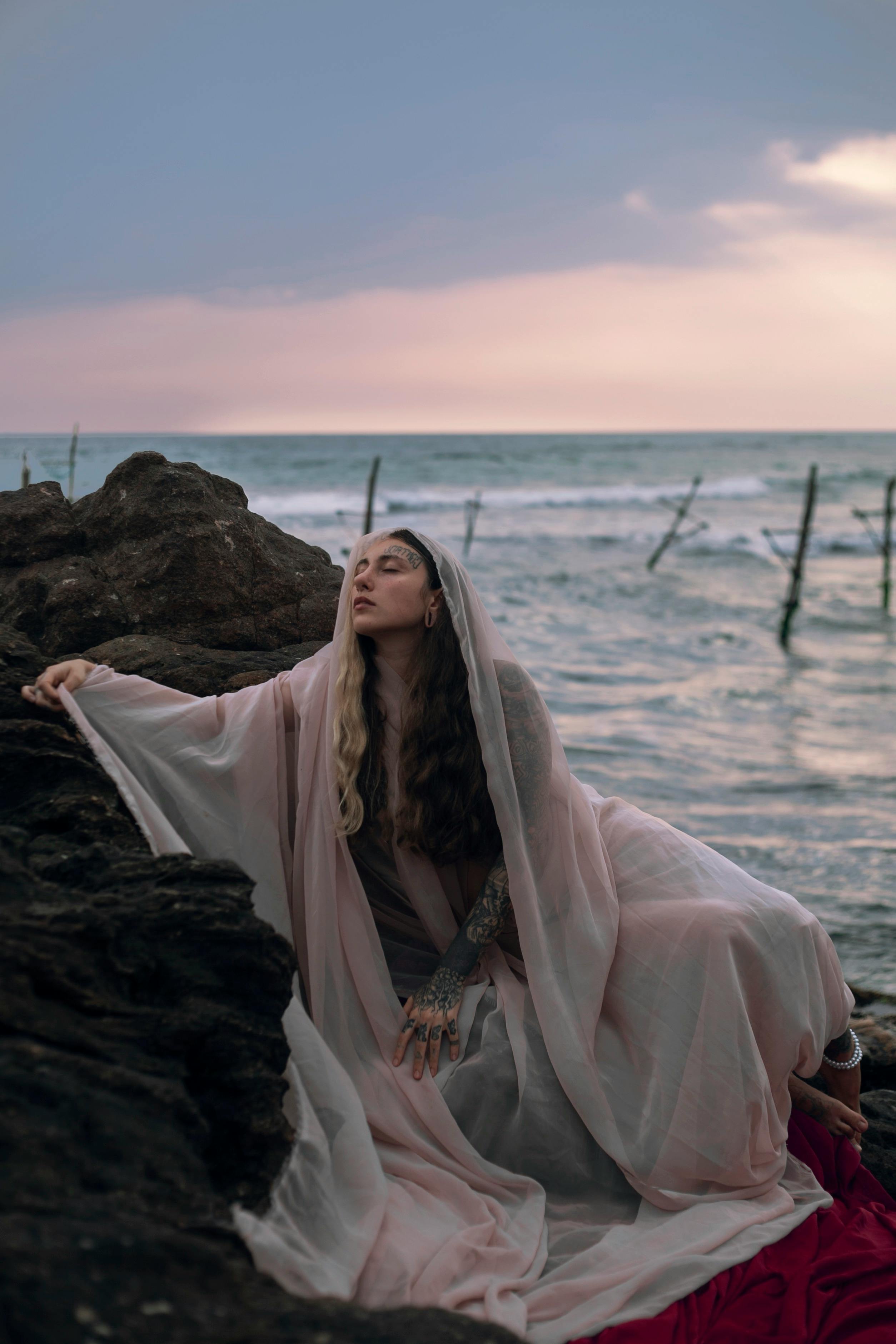 迷人的女人披着海边的透明织物，在黄昏时唤起神秘和宁静